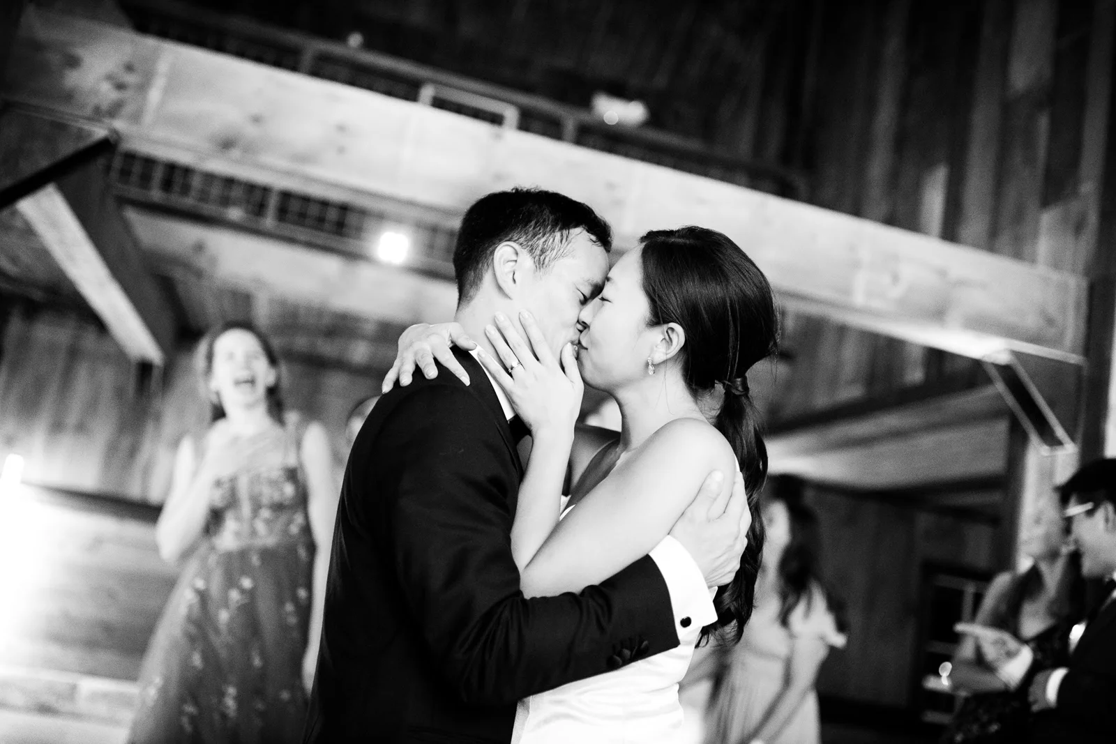 Black and white photo of a bride and groom kissing on the dance floor as a guest looks on
