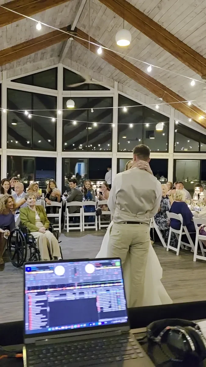 A couple sharing their first dance, seen from behind the DJ laptop, with guests seated around the room